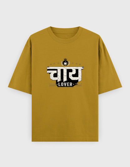 A mustard yellow short-sleeve oversized t-shirt with a distressed black and white graphic on the chest featuring the Hindi word 'सुबा' and the word 'LOVER' underneath, displayed on a light gray background.