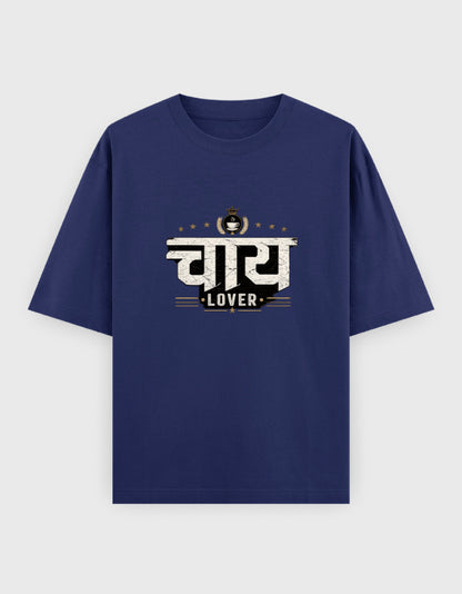 A navy blue short-sleeve oversized t-shirt with a graphic print on the chest featuring the Hindi word 'चाय 
Lover' in white and black, with the word 'LOVER' underneath and a small tea cup icon above, displayed on a light gray background.