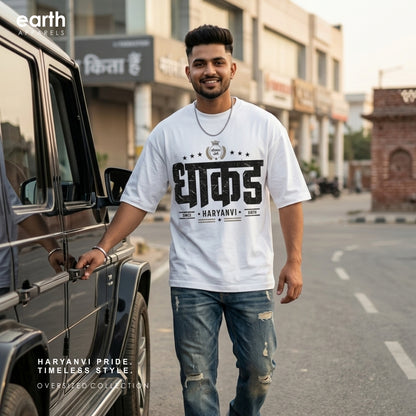 A man wearing a white graphic oversized t-shirt with Hindi text and distressed blue jeans stands next to a black SUV on a city street. The shirt features a bold black graphic print with the word 'DHAKK' and 'HARYANVI' underneath.