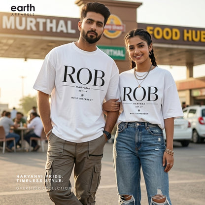 A man and a woman standing together outdoors in front of a restaurant or food hub, both wearing matching white graphic oversized T-shirts with large black text reading 'ROB' and smaller text beneath. The man wears olive cargo pants, and the woman wears blue ripped jeans.