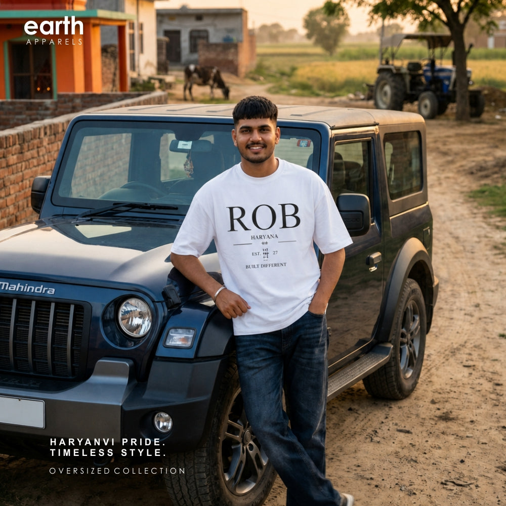 A man wearing an oversized white t-shirt with 'ROB' printed on the front, standing next to a black Mahindra Jeep on a dirt road in a rural setting. The 'earth APPAREL' logo is visible in the top left corner.